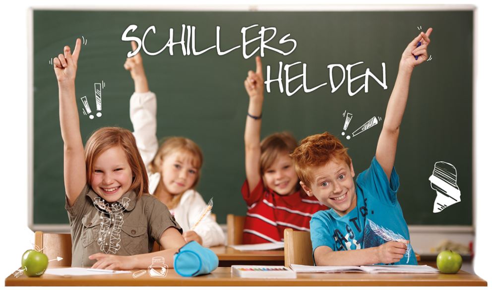 Fröhliche Schülergruppe sitzt im Klassenzimmer vor der Tafel. Auf der Tafel steht mit Kreide geschrieben, Schillers Helden.